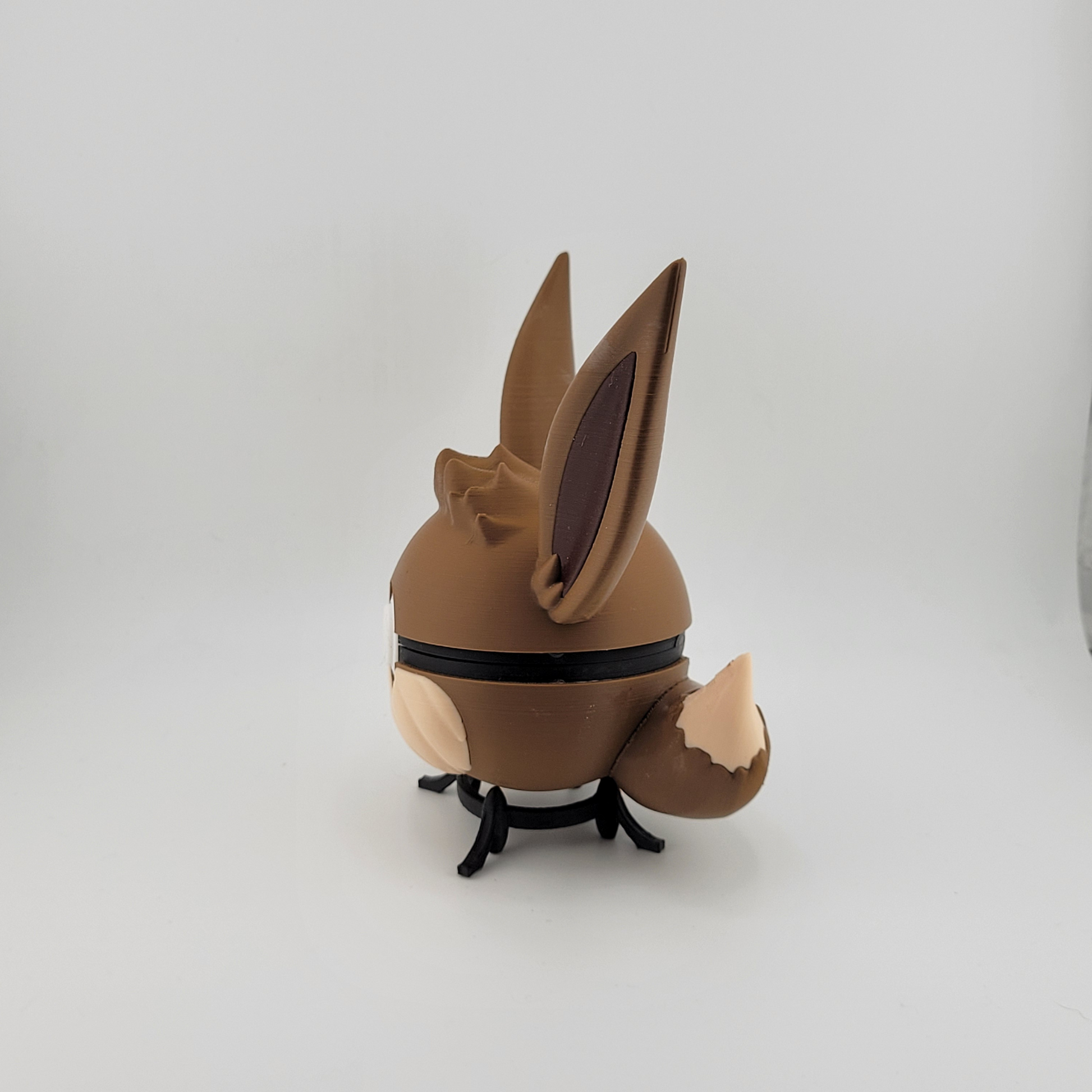 3D printed Eevee-themed Pokéball with brown and cream colors, featuring Eevee ears and tail design, displayed on a stand.