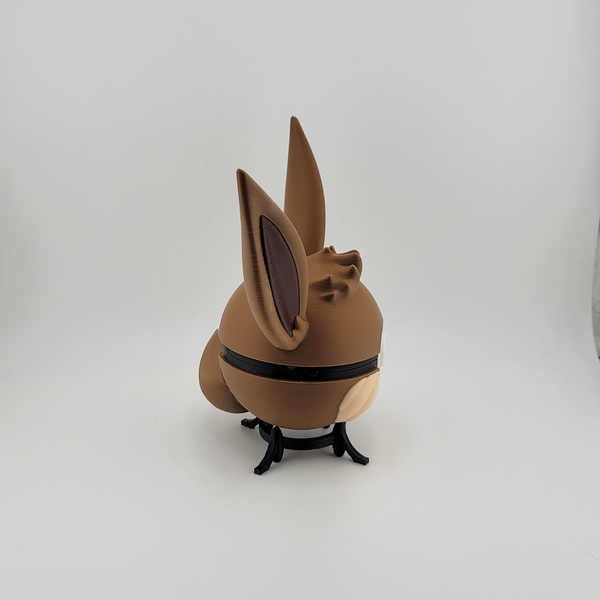 3D printed Eevee-themed Pokeball with brown and cream colors, featuring Eevee's ears and face, displayed on a black stand against a white background.