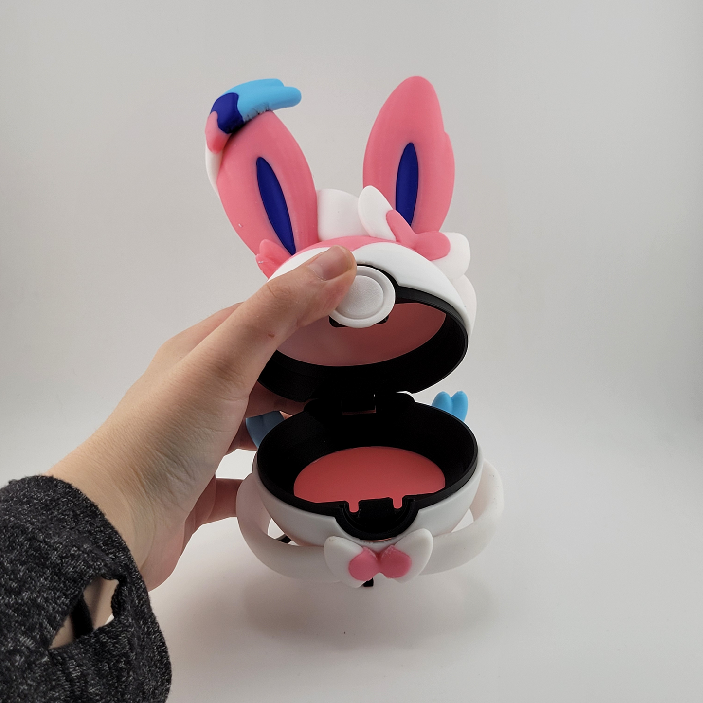 Hand holding an open Sylveon-themed Pokéball with pastel pink, blue, and white colors and ribbon motifs, designed for storing Pokémon figures or collectibles.