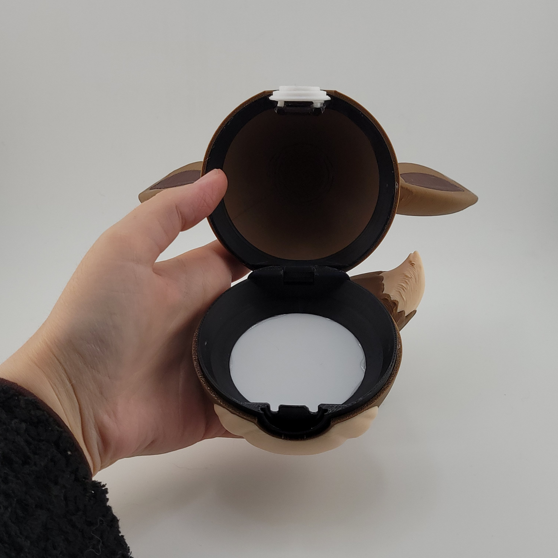Hand holding an open Eevee-themed Pokeball designed with brown and cream colors and Eevee ears and tail details.