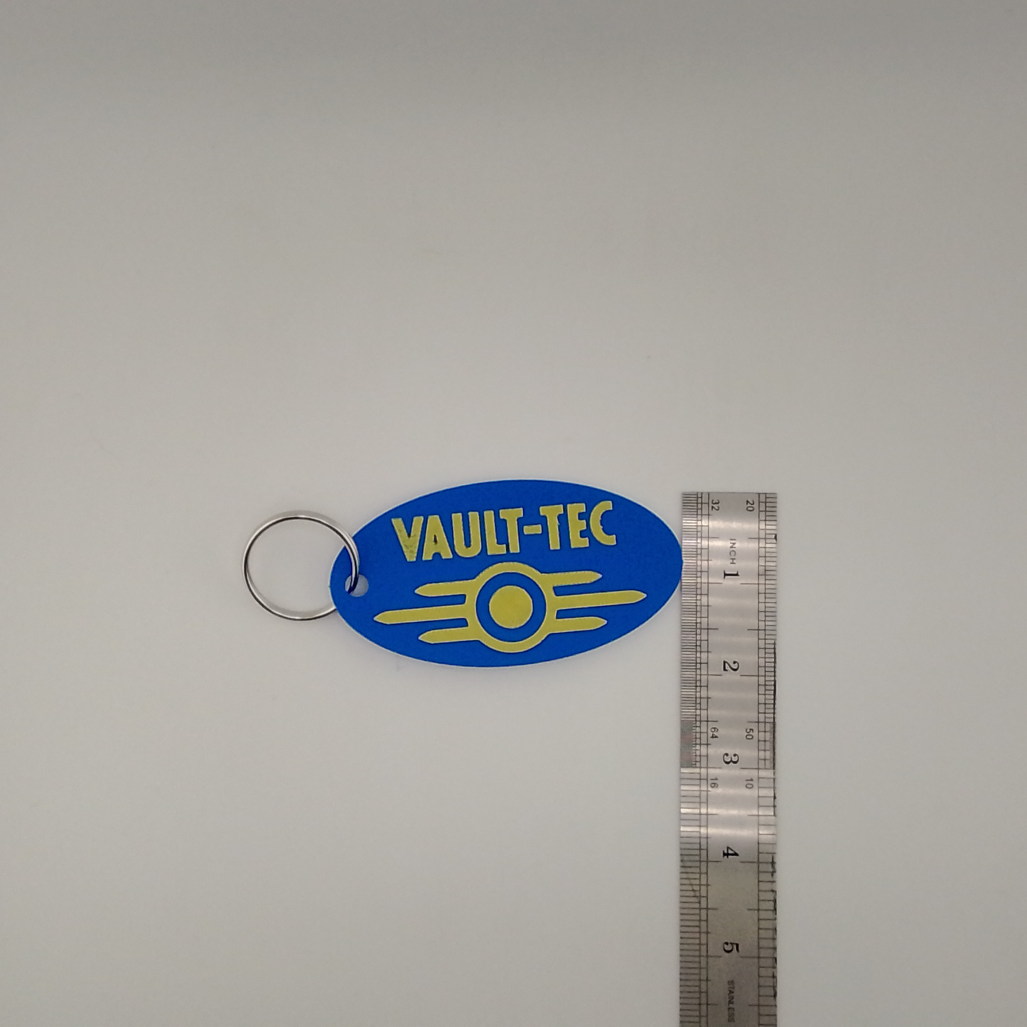 Blue 'Vault-Tec' keychain with yellow text on a gray background with a ruler for scale.