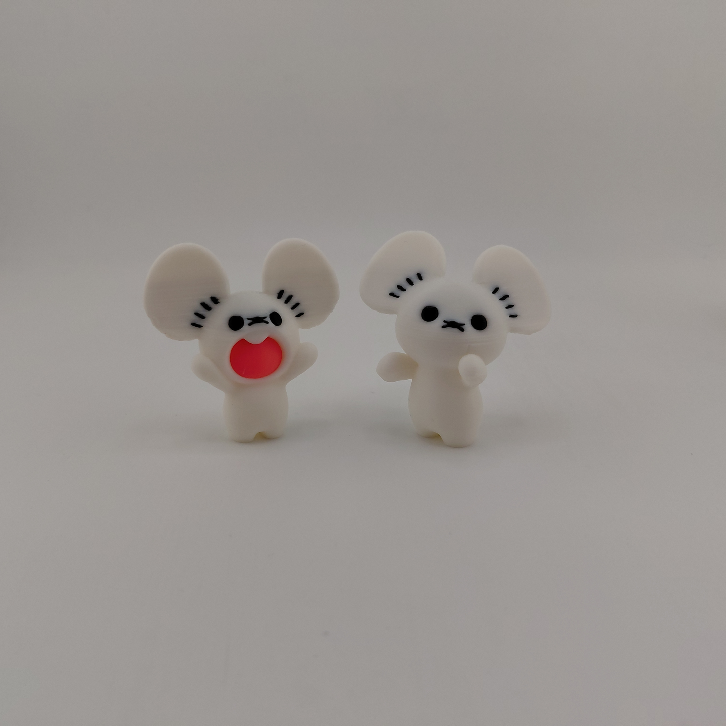 Two small white cartoon mouse figurines standing side by side on a plain light gray background. One has a red heart-shaped mouth and the other has a simple black face.