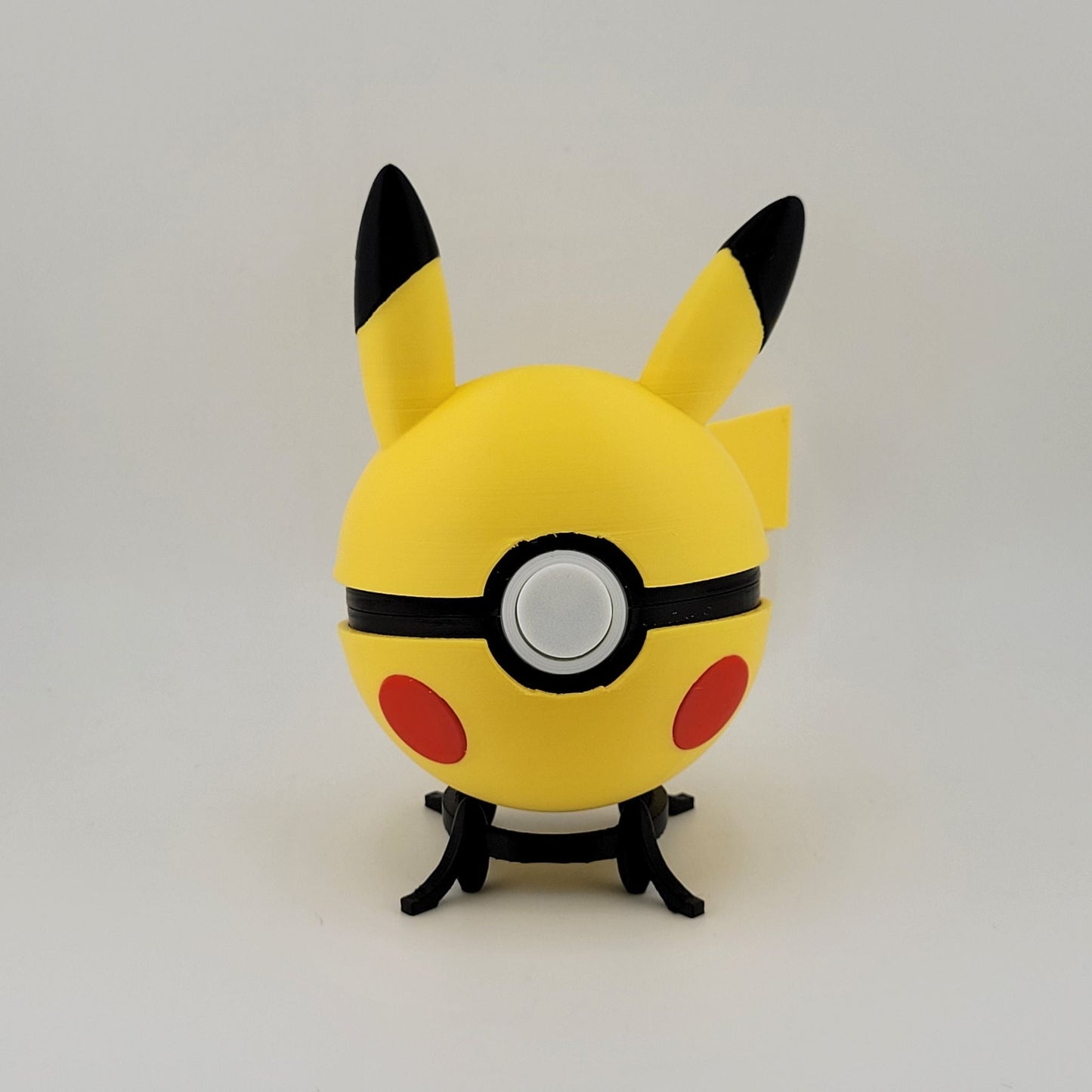 3D printed Pikachu-themed Pokeball with yellow body, black-tipped ears, and red cheeks on a black stand