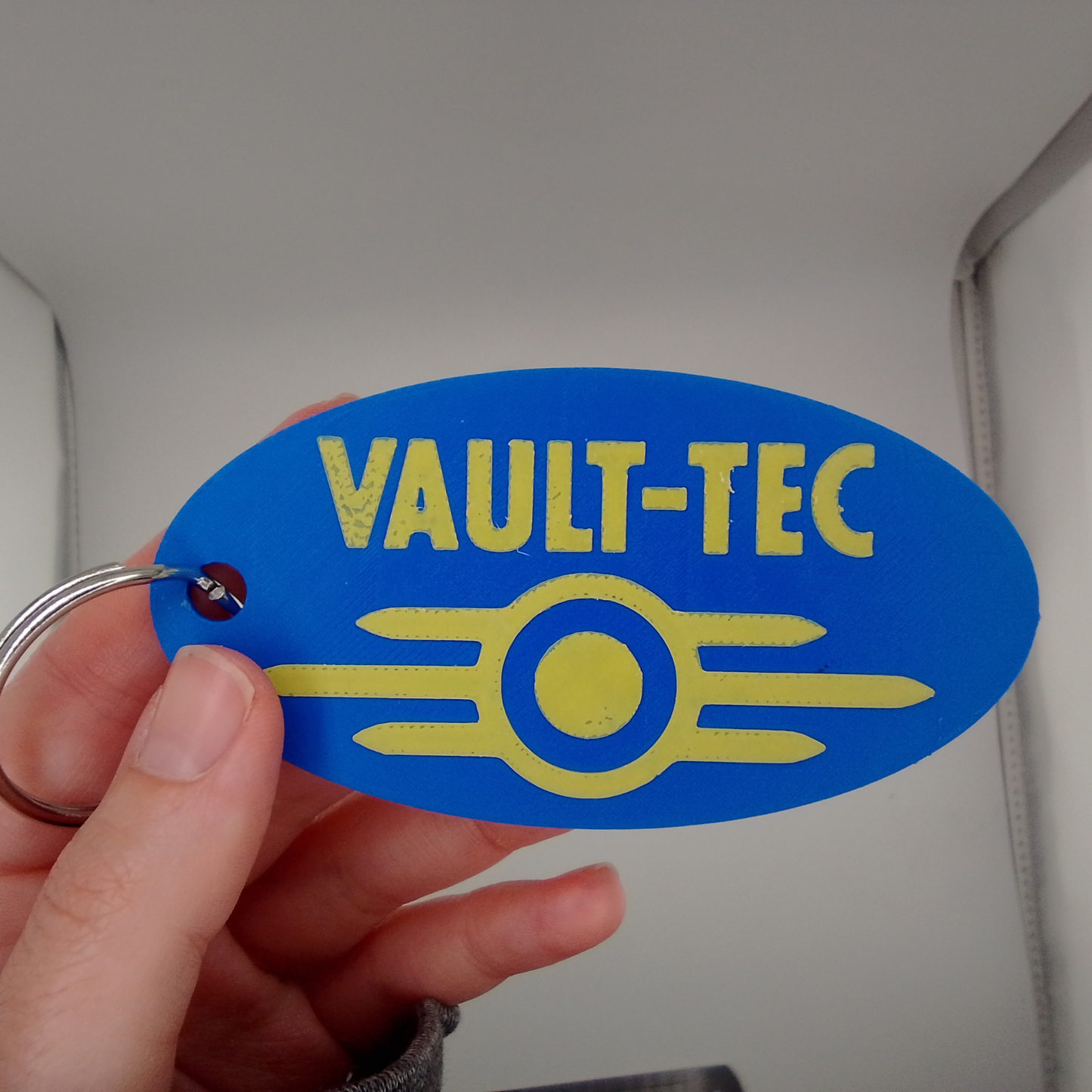 Blue 'VAULT-TEC' keychain tag held by a hand against a neutral background