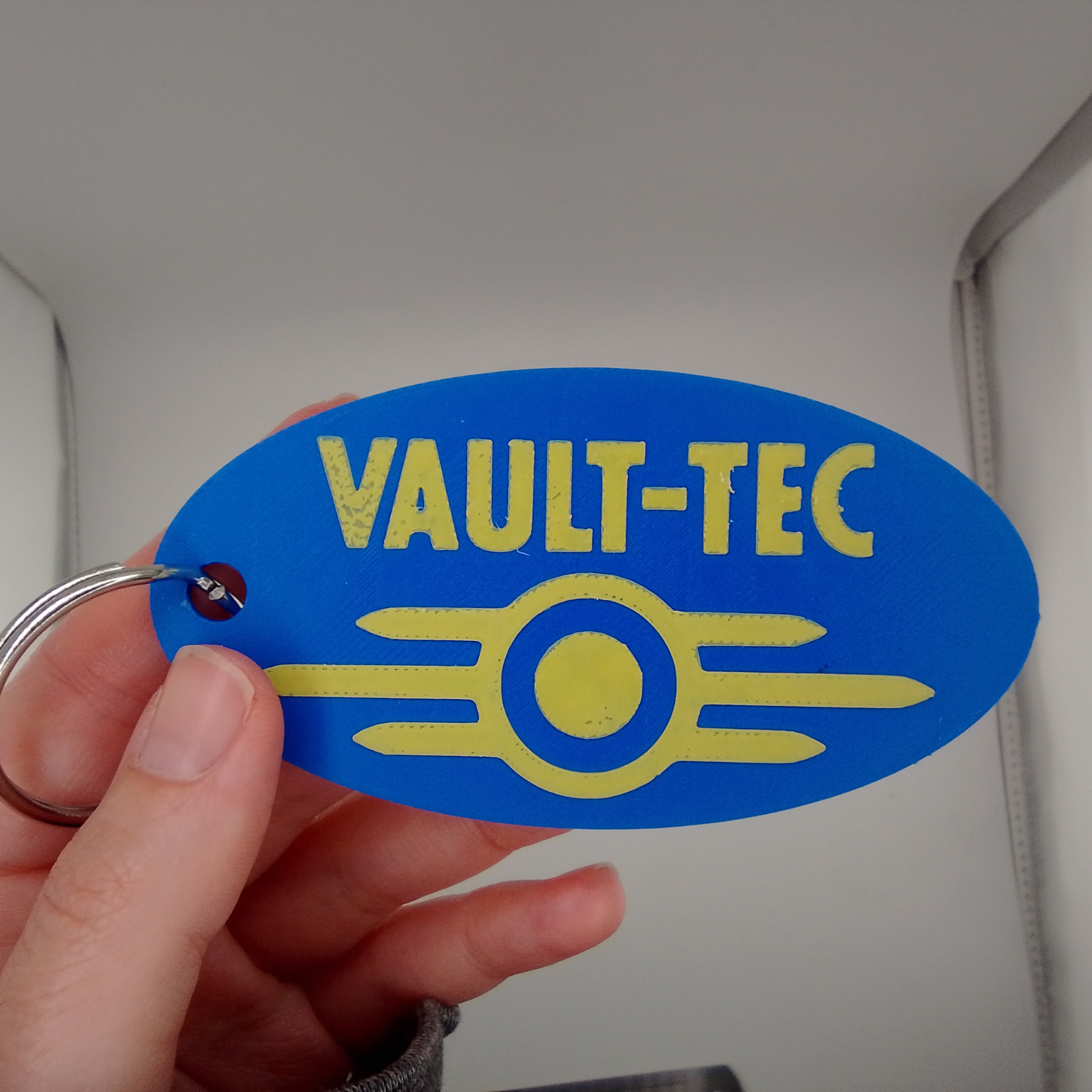 Blue 'VAULT-TEC' keychain tag held by a hand against a neutral background