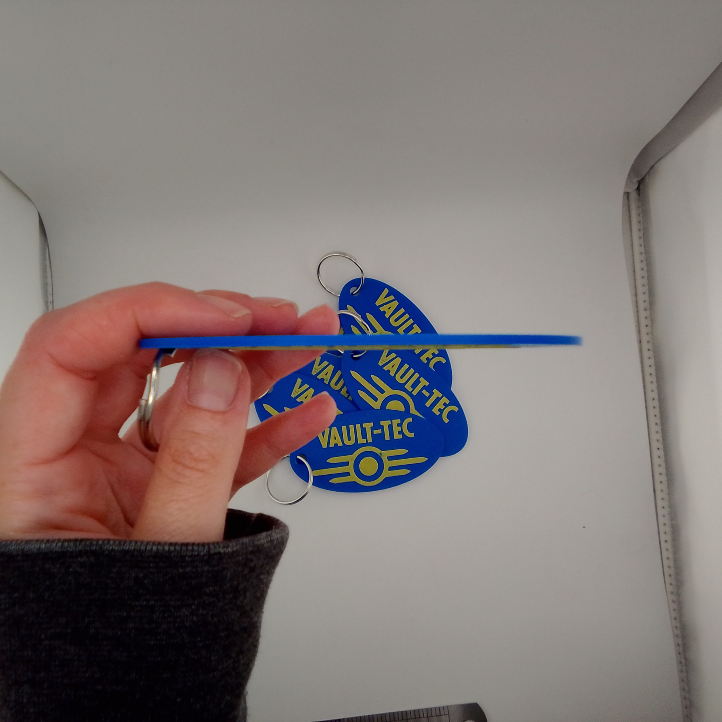 Hand holding a blue stick with two 'Vault-Tec' keychains attached against a plain background