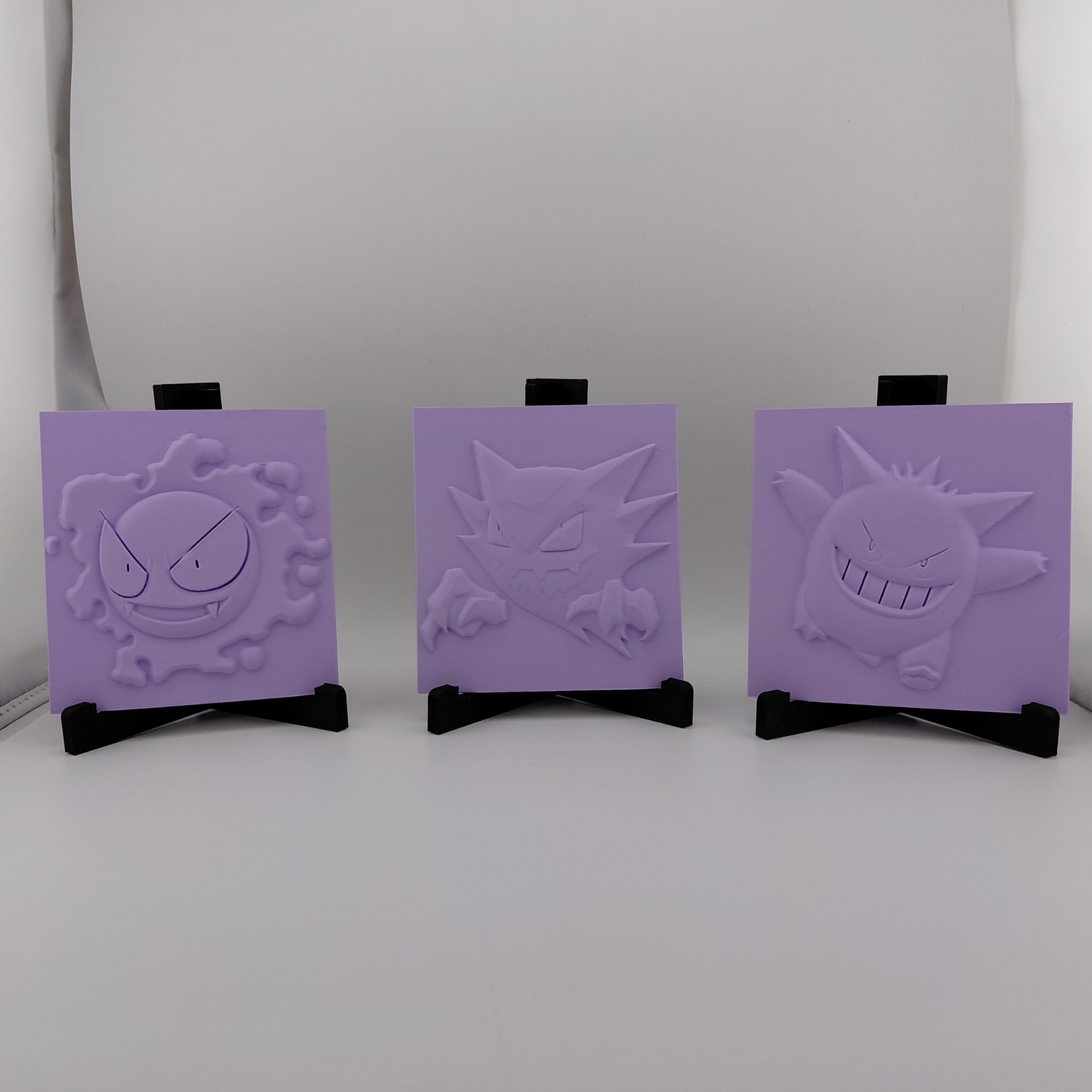 Set of three lavender 3D printed relief plaques featuring Gastly, Haunter, and Gengar inspired ghost monsters displayed on black stands against a white background. Each square plaque has raised detailed artwork and comes with its own stand for desk, shelf, or gaming room decor. Available individually or as a matching set for collectors and fans of spooky monster-inspired decor.