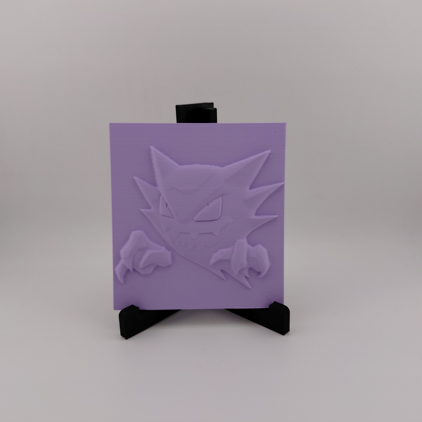 Set of three lavender 3D printed relief plaques featuring Gastly, Haunter, and Gengar inspired ghost monsters displayed on black stands against a white background. Each square plaque has raised detailed artwork and comes with its own stand for desk, shelf, or gaming room decor. Available individually or as a matching set for collectors and fans of spooky monster-inspired decor.