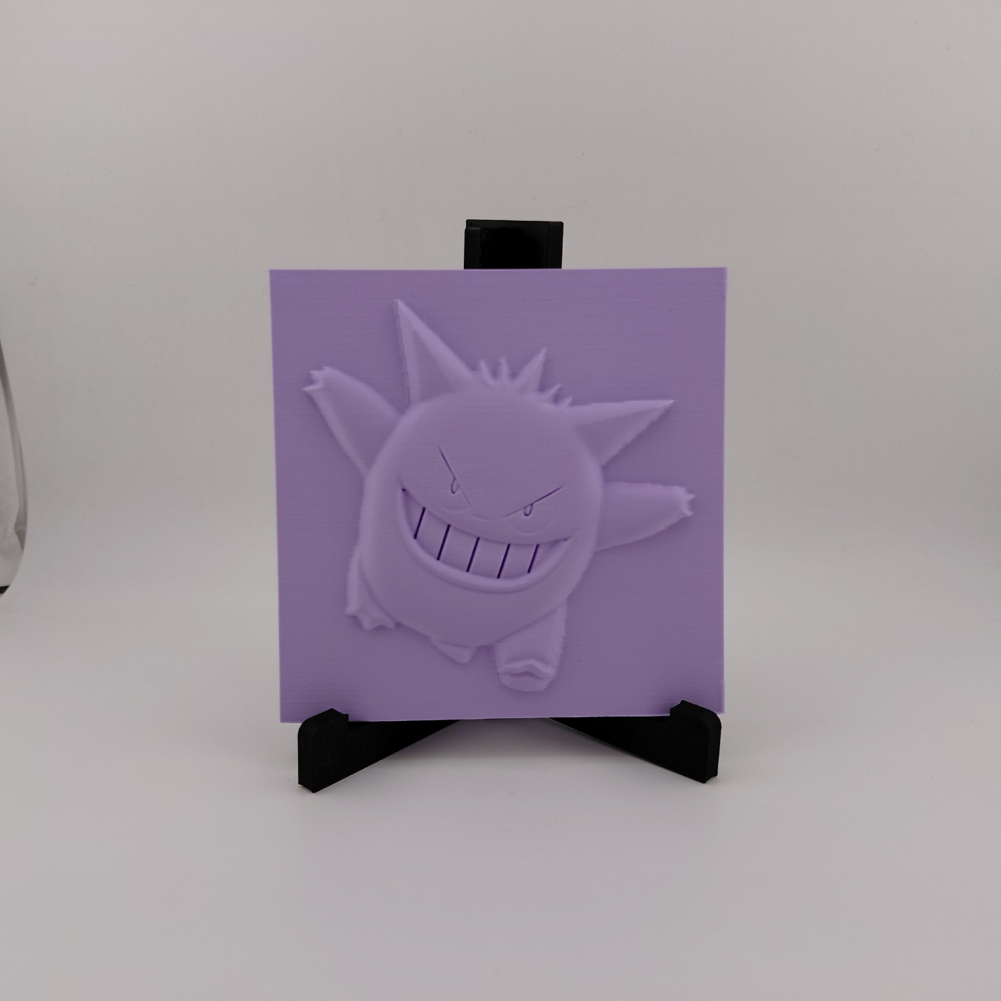 Set of three lavender 3D printed relief plaques featuring Gastly, Haunter, and Gengar inspired ghost monsters displayed on black stands against a white background. Each square plaque has raised detailed artwork and comes with its own stand for desk, shelf, or gaming room decor. Available individually or as a matching set for collectors and fans of spooky monster-inspired decor.