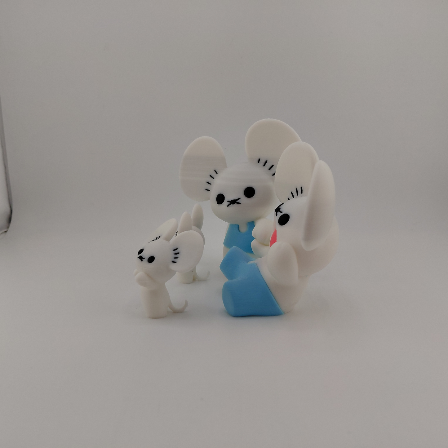 Three small white mouse figurines with black facial features and one wearing a blue outfit, photographed on a plain light gray background.