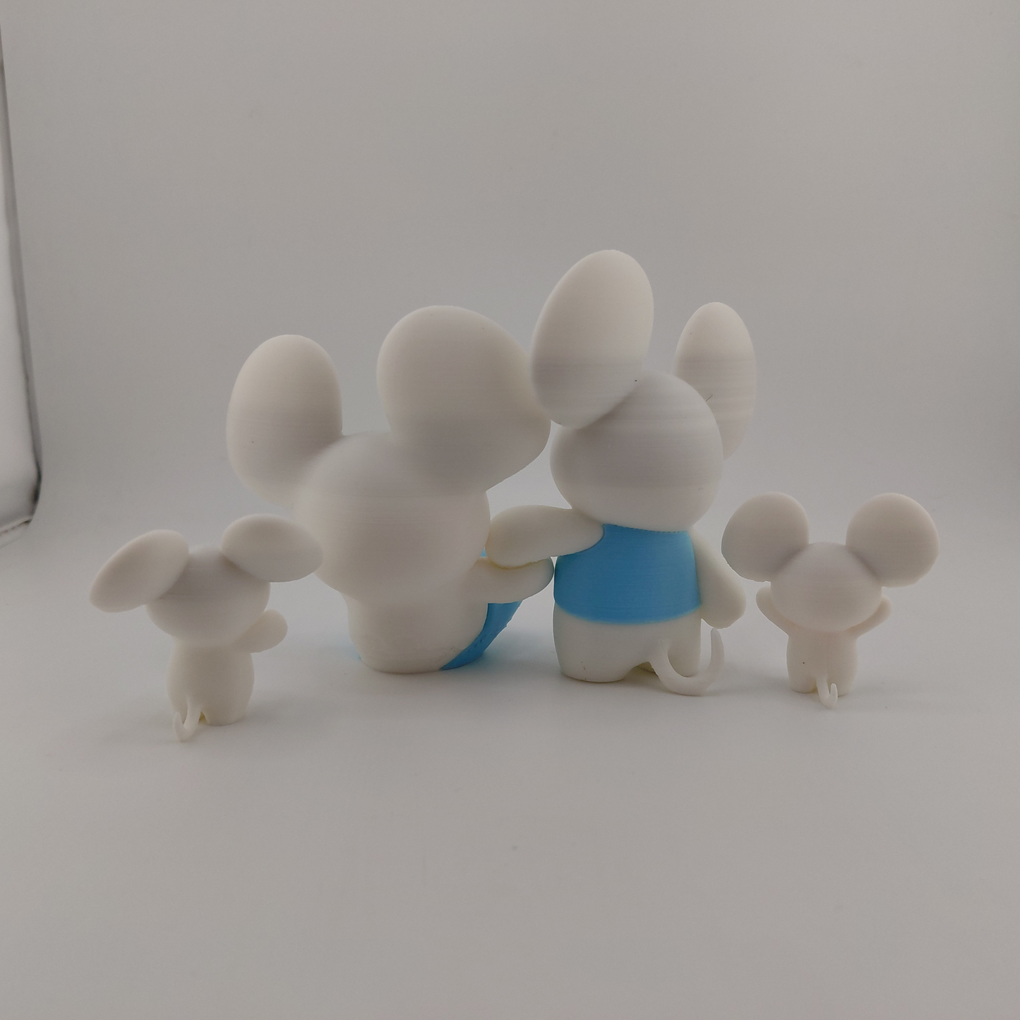 Four white mouse figurines with large round ears, posed together on a plain light gray background. The two larger figures in the center wear light blue clothing, while two smaller figures stand on either side.
