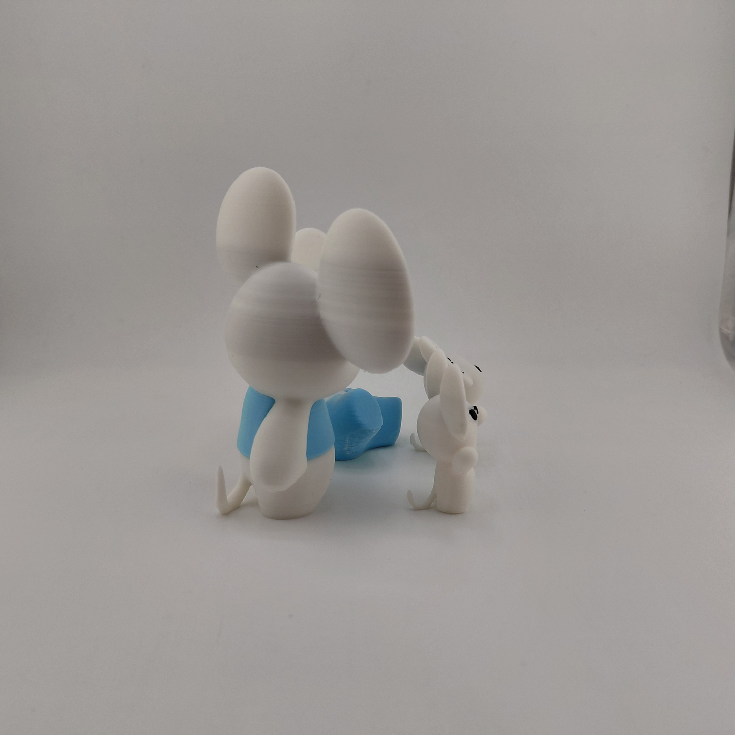 A white plastic figurine of a mouse-like character with large ears, wearing a blue garment, posed next to a small white animal figure on a plain light gray background.