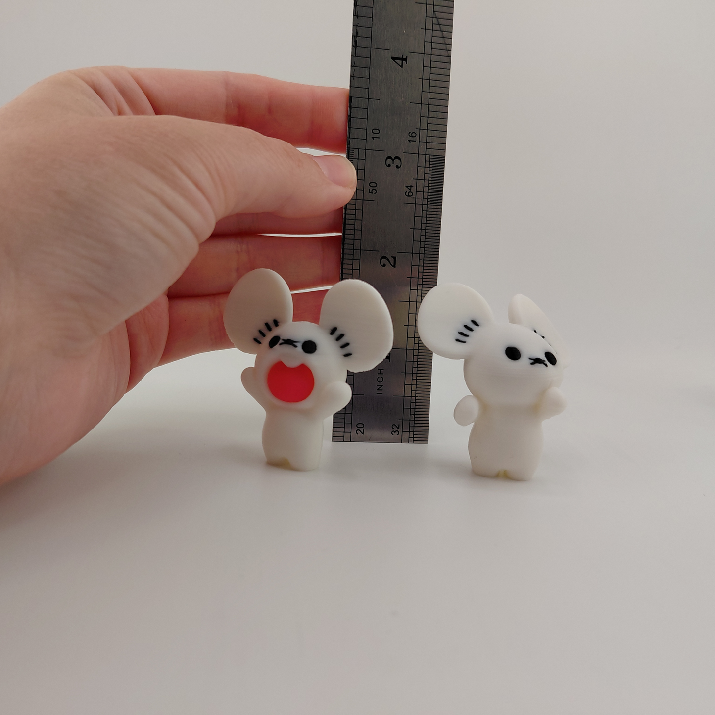 Two small white mouse figurines with black facial features and one red mouth piece, shown next to a metal ruler and held by a human hand to demonstrate scale.
