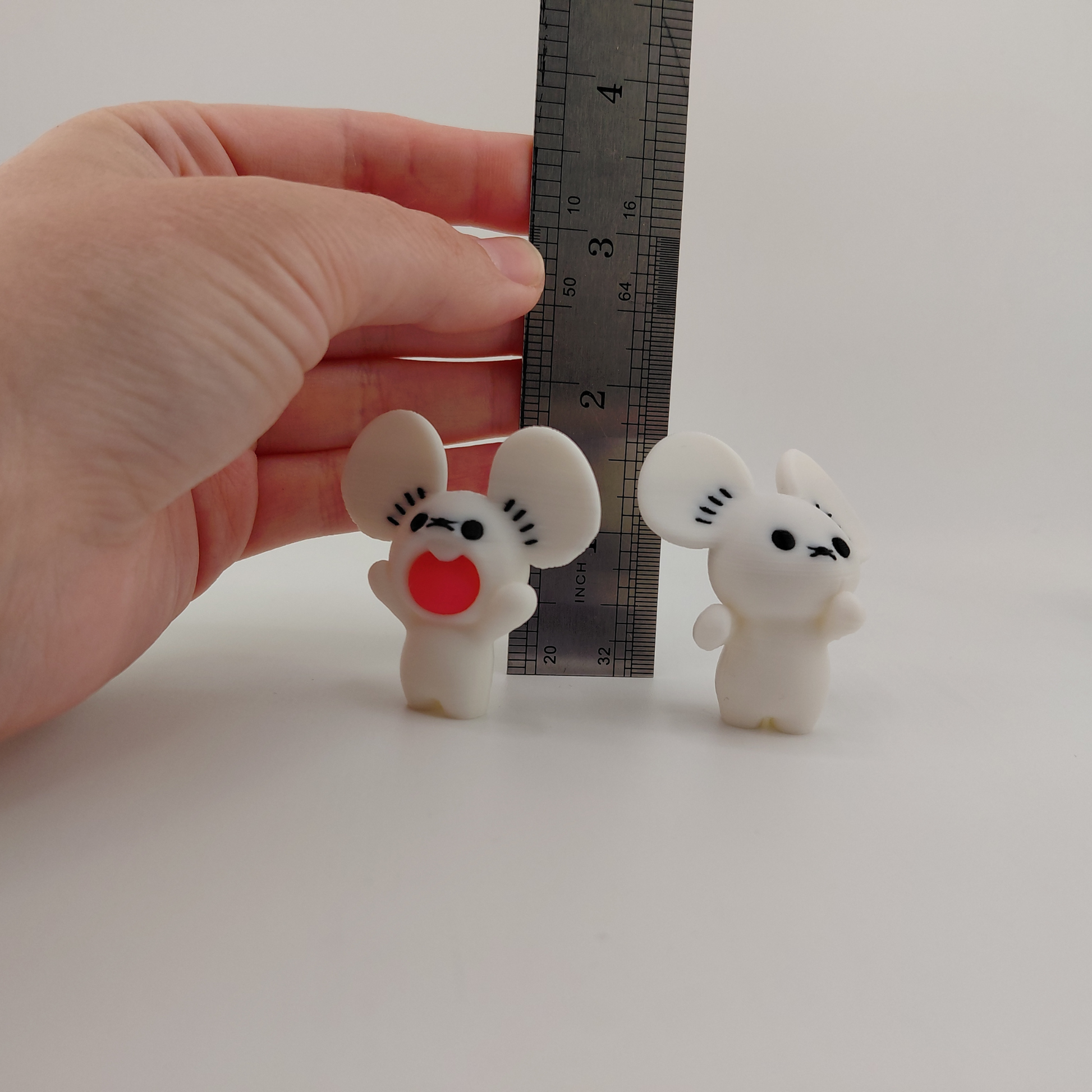 Two small white mouse figurines with black facial features and one red mouth piece, shown next to a metal ruler and held by a human hand to demonstrate scale.