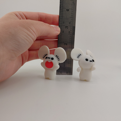 Two small white mouse figurines with black facial features and one red mouth piece, shown next to a metal ruler and held by a human hand to demonstrate scale.