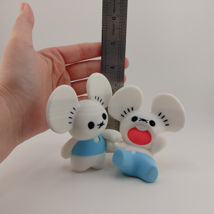 Two small white mouse figurines with black facial features, one wearing a light blue shirt and the other blue pants, shown next to a metal ruler and held by a person's hand to indicate size.