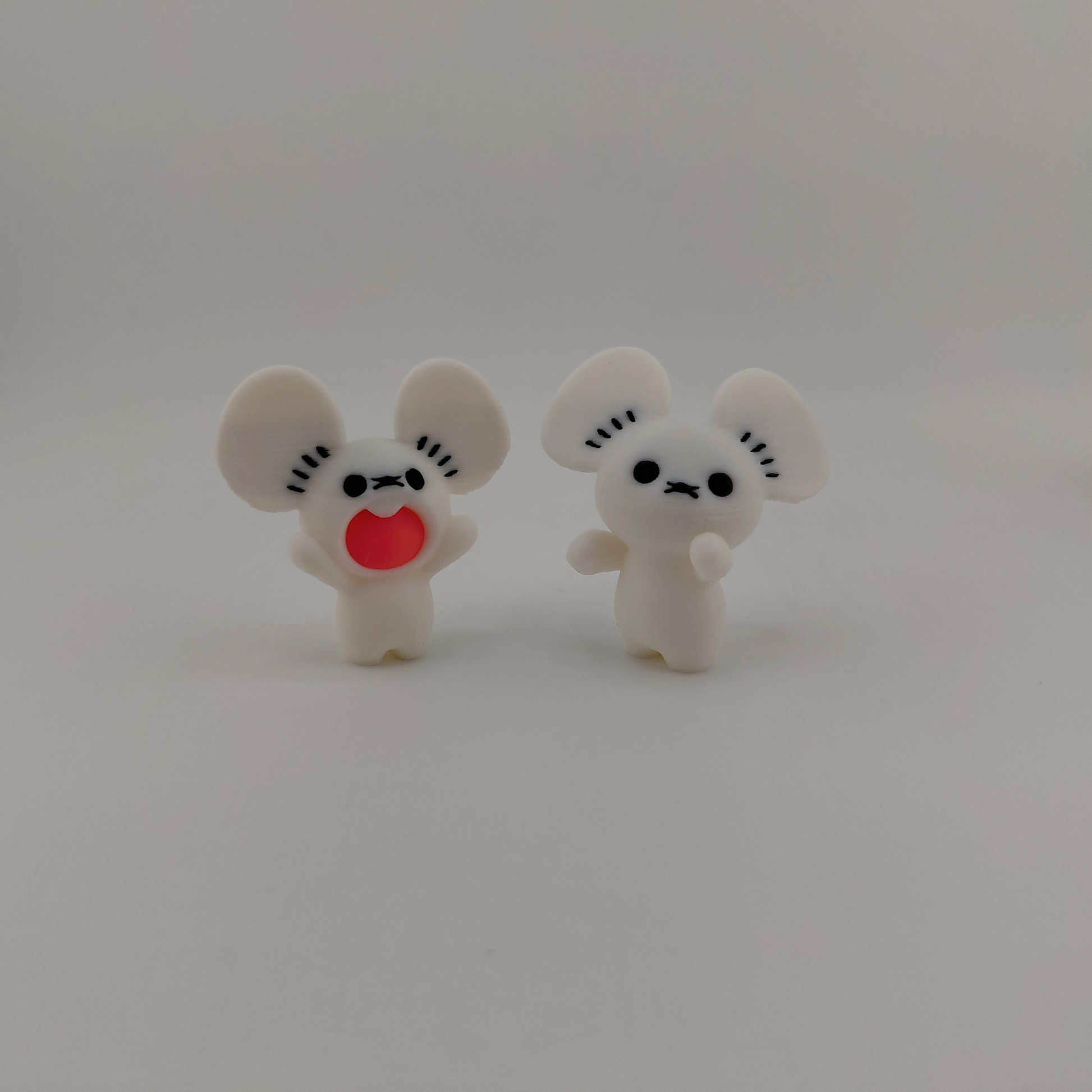 Two small white cartoon mouse figurines standing side by side on a plain light gray background. One has a red heart-shaped mouth and the other has a simple black face.